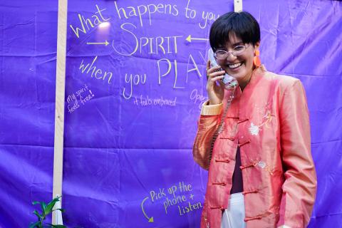 An Asian woman poses with a corded phone in front of a backdrop that reads: "What happens to your spirit when you play?"