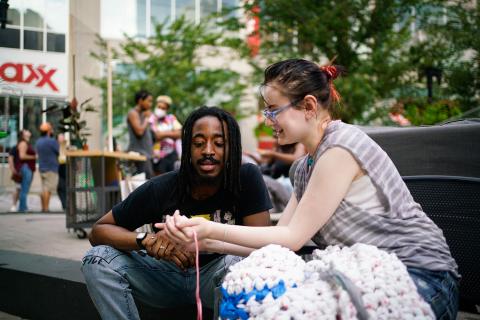 A white woman and a Black man, with long braids, hold yarn together.