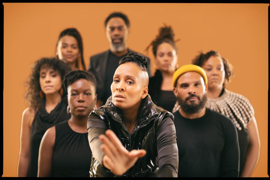 In front of a brown backdrop, eight folks of color pose in black outfits. A woman in the foreground holds her hand up.