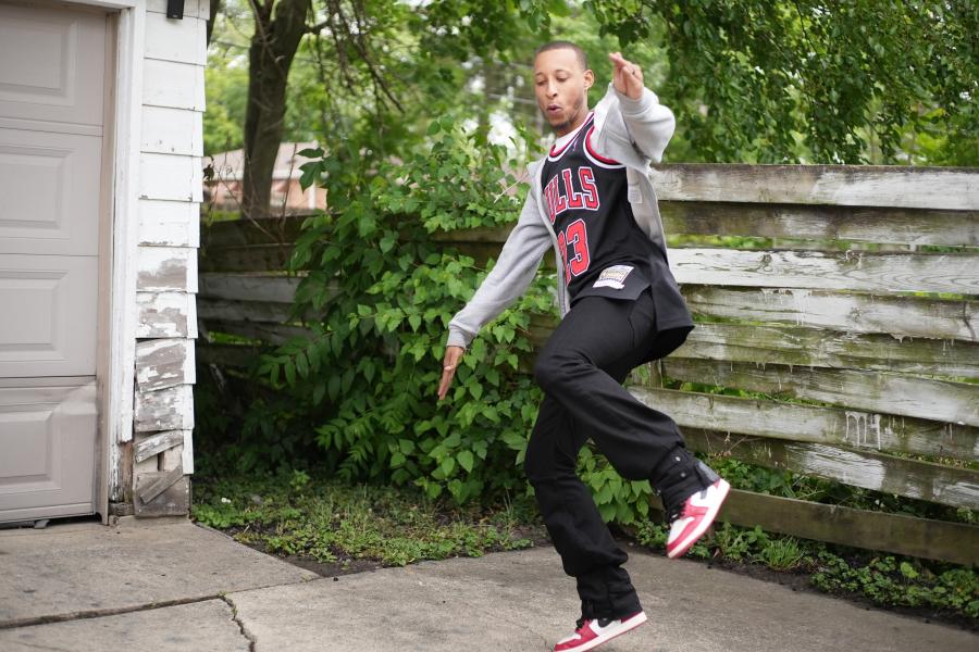 By a garage, a young Black man in a Chicago Bulls basketball jersey dances.