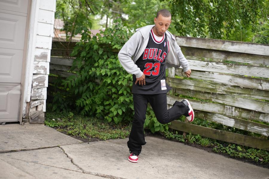 By a garage, a young Black man in a Chicago Bulls basketball jersey dances.