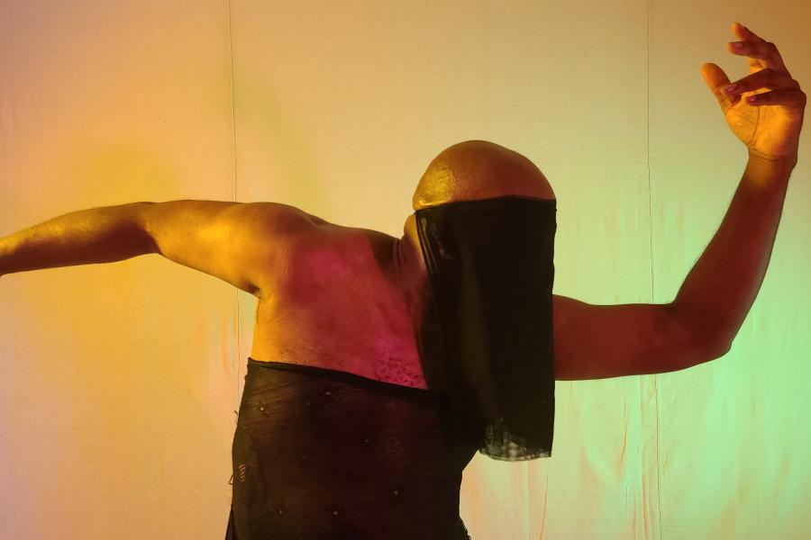 A Black person holds their arms out to their sides, with a black cloth over just their face, revealing their bald head.