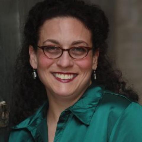 Woman in glasses with her hair pulled back, smiles in her teal button down
