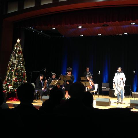 Image of a woman singing into a microphone from a stage, accompanied by musicians. A Christmas tree is on the left.