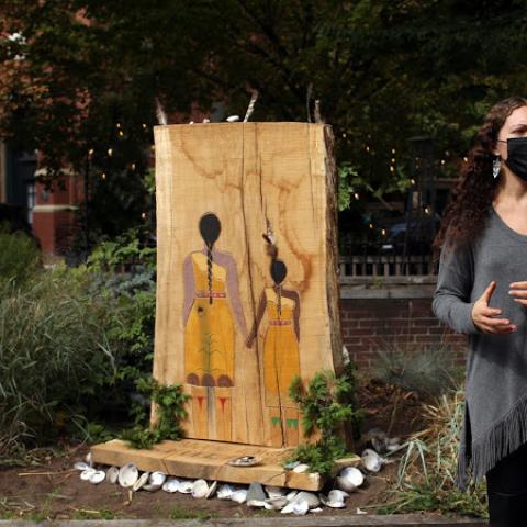 Lily, in a face mask, speaks next to an installation, two Native American women painted on a shaved tree trunk.
