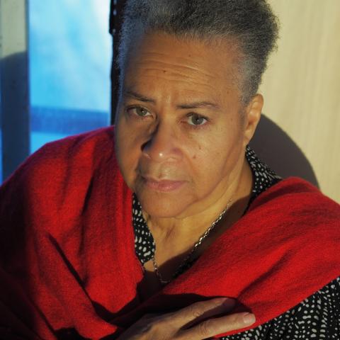 Linda has a red scarf wrapped around her. She is a Black woman with short grey and white hair. She is spotlit by a warm lamp.