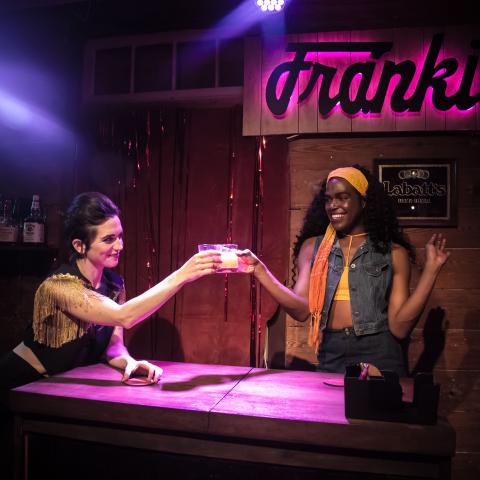 Two women stand by a bar that says "Frank's" in neon behind them,