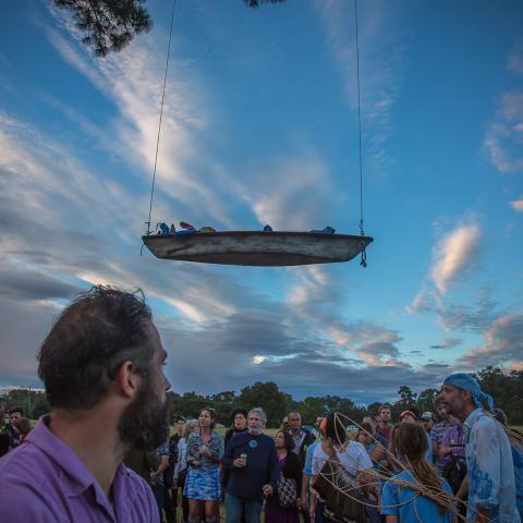 A boat hangs in the air as if it is floating over a group of people, outside.