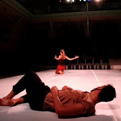 In the foreground, a male dancers lays on the floor watching a female dancer, on her feet in the background.