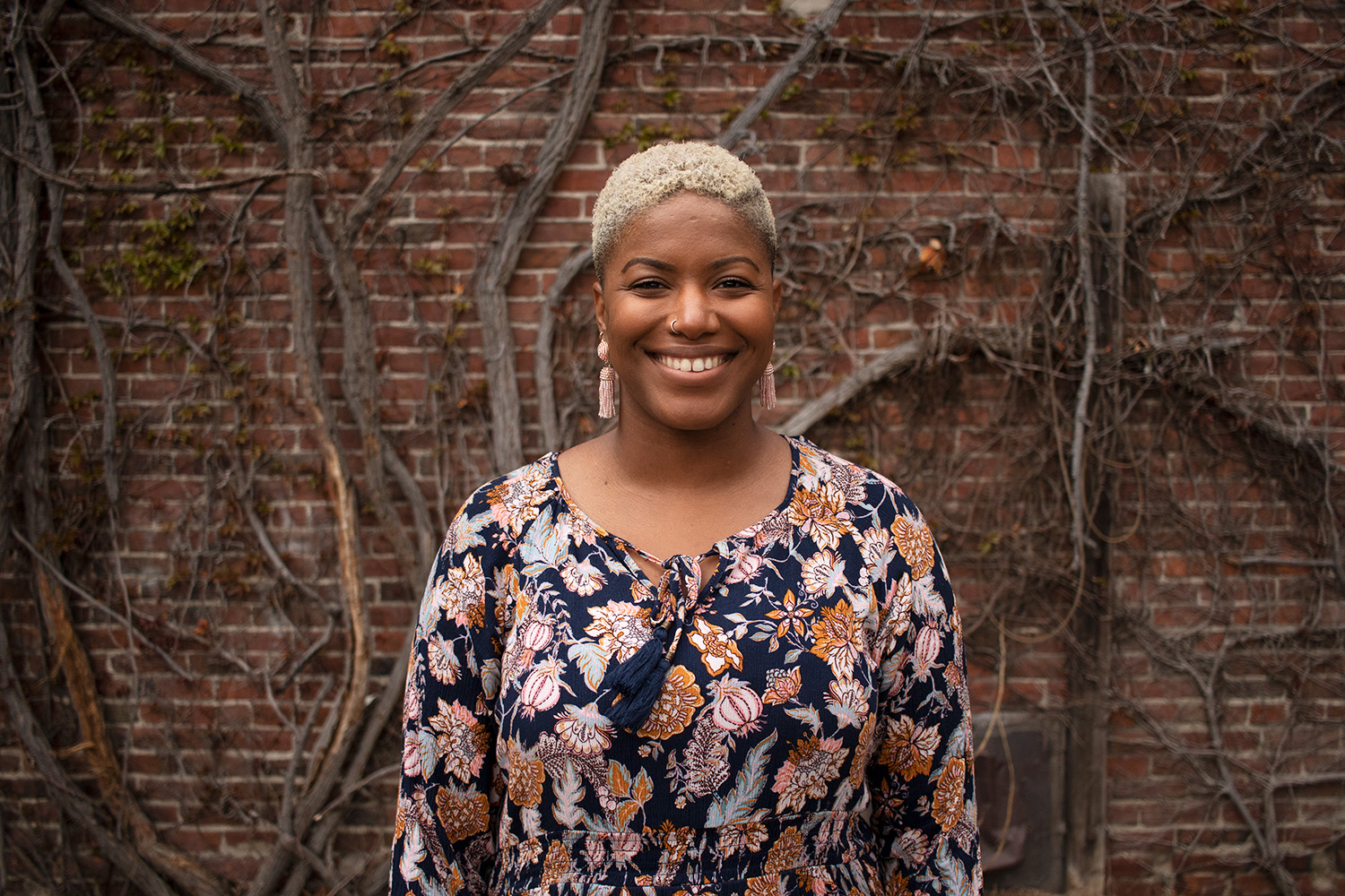 Christa is a woman of color. She has short bleach blonde hair and wears a floral dress. She poses in front of a brick building with vines.