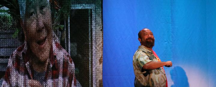 Next to a projection of an older woman, a spotlit man stands and turns to the audience.