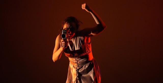 In dramatic red and yellow lighting, a light-skinned woman holds her fist up and looks through a super 8 camera lens.