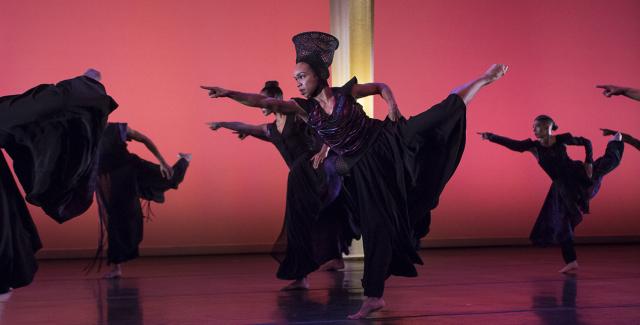A dancer, in an ensemble of dancers, lifts her leg back and up and points forward.
