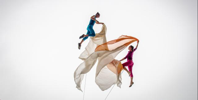 In a white void, two dancers (in brightly colored costumes) dance with orange and beige fabric.