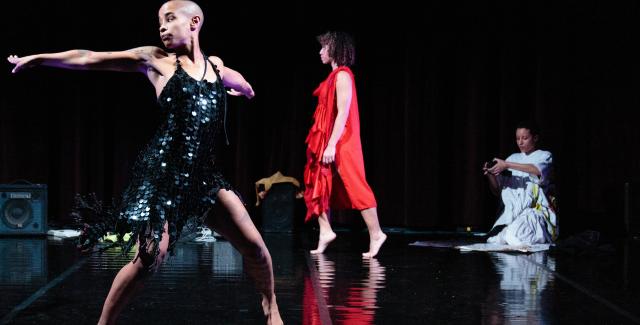 On a stage, a person in a Black flapper dress dances while a person in a red romper hops down and another person in a white dress kneels.