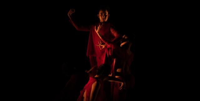 A dimly lit person, holds their hands up to their right and wears a long red cape.