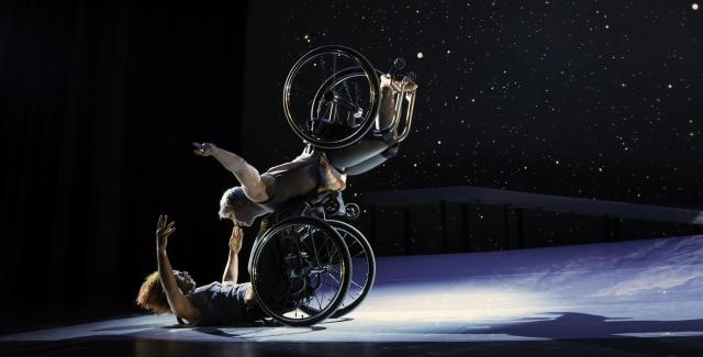 Two wheelchaired dancers in a starry setting. One is on their back on the ground and the other is somehow leaning on top of them without either leaving their chairs.