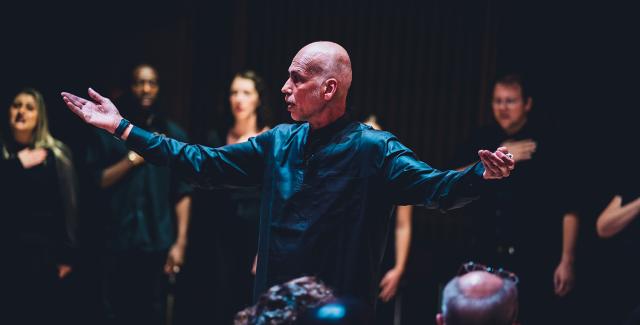 A bald man holds his arms out, in front of a group wearing all black.