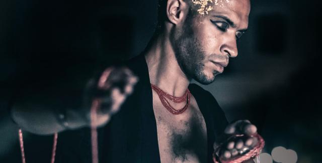 A young, Brown man holds triangle instruments and wears gold leaf on his bald head.