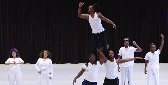 Seven Black performers on a white stage. One man, in a wife beater, is on the shoulders of two of his peers and looks out to the left.