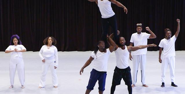 Seven Black performers on a white stage. One man, in a wife beater, is on the shoulders of two of his peers and looks out to the left.