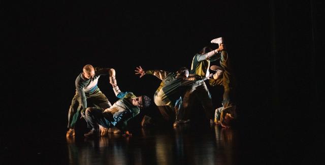 In a dark studio space with spotlights like flashlights, dancers appear to play fight.