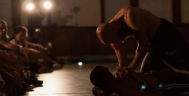 In very low light, one shirtless man is on top of another. An audience sits on the floor beside them.