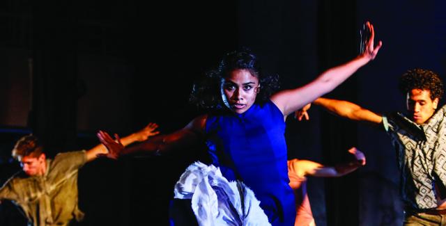 A woman in blue dances in the foreground, with male dances dimly lit in the background.