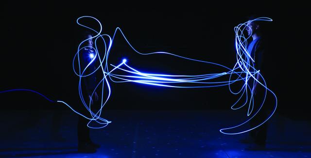 A man and a woman stand still while someone runs around with a small light to create shapes around them during a long exposure.