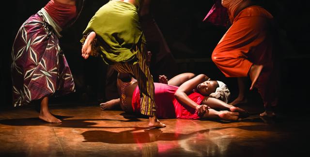 Five women in bright colors dance around two women embracing on the floor.