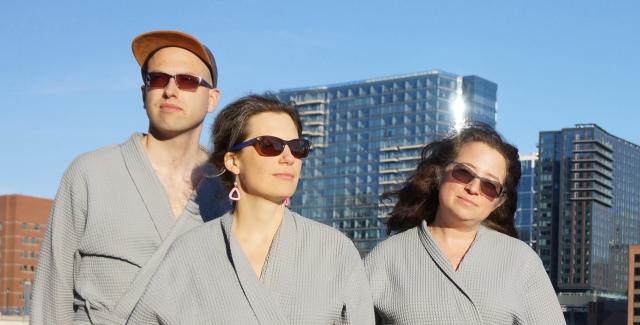 A man and two women in robes and sunglasses pose by the waterfront.