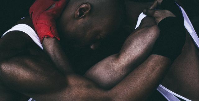 Two Black men in wrestling singlets and gloves hold each other in a head lock that looks almost like a hug.