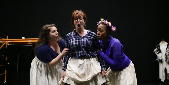 On a stage, three women in aprons argue.