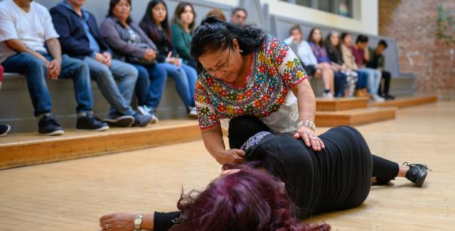 An audience watches as a woman performs assisting another woman, who lies on the ground.