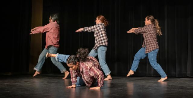 Four dancers, on a black stage with black curtains, wear denim and flannel. Three face left and throw their arms that way; another is on the ground kicking in the same direction.
