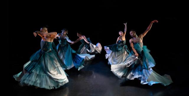 a photo features five trans, queer, and gender-expansive Sean Dorsey Dance dancers wearing elaborate floor-length blue and silver strapless ball gowns, dancing in a circle on stage. Each dancer is twirling around in a circle and has their arms out in different expressions of grace and longing. 