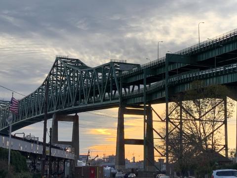 A view of the Tobin bridge from below with a cloudy sky with a bit of sun in the background