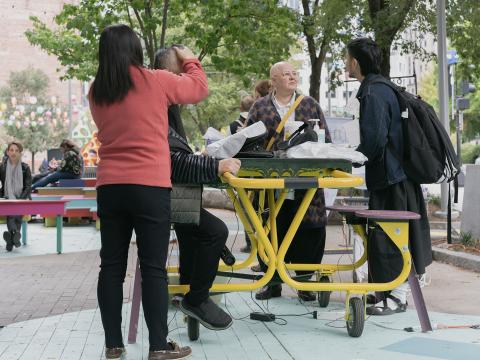 Outside, in a park with a yellow table and benches, four folks, most of them Asian folks, gather. One woman rubs the head and back of a man. A man speaks to a woman across the table from them.