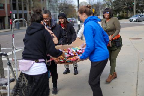 Outside on a city street, six women carry cardboard with colorful parts laid on top of it.