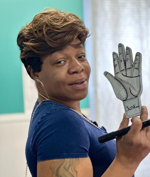 A Black woman holds up an painted, cutout of a palm with the name "Justin" at the bottom.