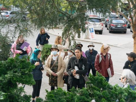 Outside, between some trees and bushes, folks wait. Some wear masks. 