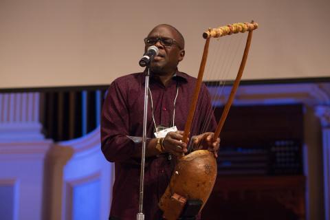 artist Samite Mulondo plays a litungu, a seven stringed instrument, and sings into a standing mic at the 2018 Idea Swap on the Great Hall stage at Mechanics Hall. Samite's eyes are closed as he performs. Blue lighting on the stage and a blank projector screen are seen in the background.
