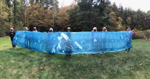 In a field, seven women hold up a blue banner.