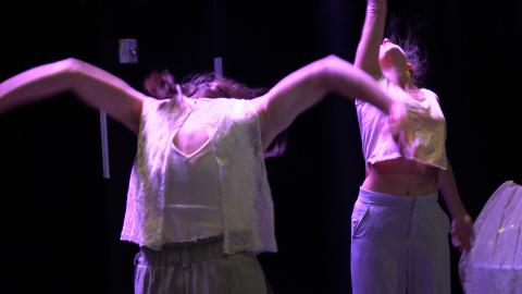 On a stage, with purple lighting, two women dance and there is motion blur.