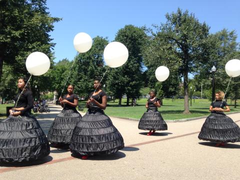 Five women of color in black dresses, holding white balloons by white ribbons, walk through a park.
