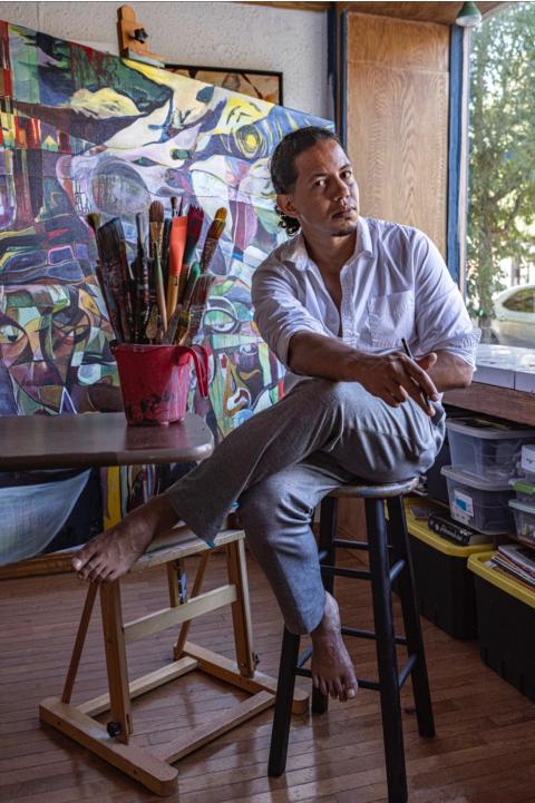 A brown man poses on a stool. Next to him are large paint brushes in a bucket and a a painting on an easel the size of him.