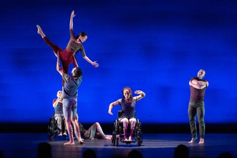A dancer in a wheel chair and standing dancer hold another dancer in the air. Another dancer in a wheel chair holds her arms in a circular formation. Another dancer stands and crosses his arms.