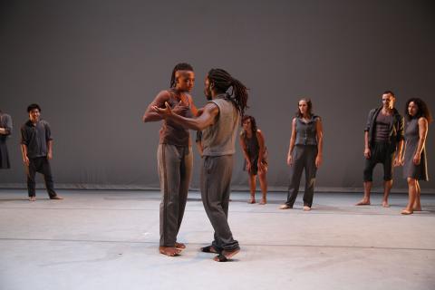 Dancers pose and wear all grey in a grey space with a white floor. Two Black folks in the center hold hands like they're waltzing.