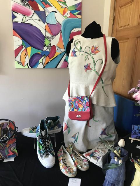 Display of artwork in a studio; colorful, graphic designs on canvas as well as a dress form, purse, and sneakers.