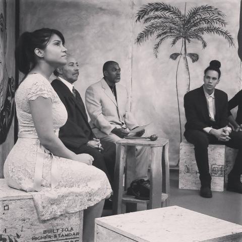 a group of a Latinx performers sit on a stage staring dramatically into the distance - like a sepia tone telenovela
